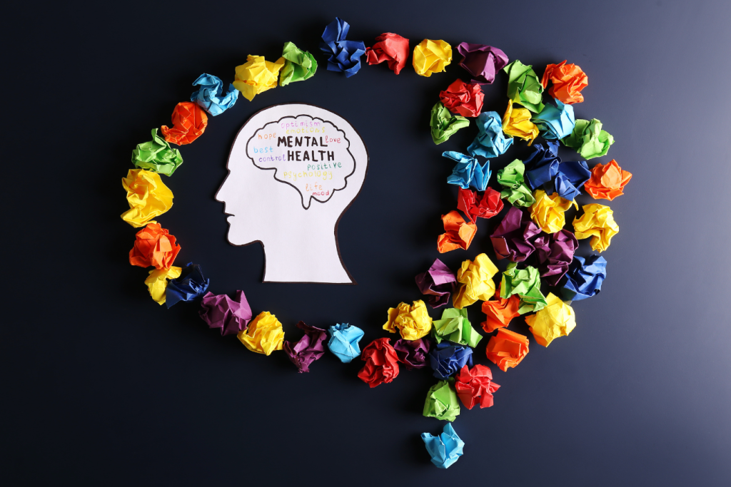 A cardboard cut-out of a head surrounded by scrunched up paper in various colours that forms the shape of a speech bubble. Inside the head is an outline of a brain in which are the words Mental Health.