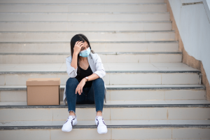 A woman in a facemask sits on some steps, her head is in her hands and a box sits beside her